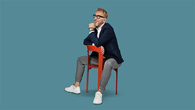 Mann mit modischer Brille und Sakko sitzt auf einem roten Stuhl