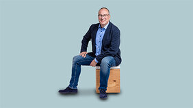 Mann in Jeans und Sakko sitzt auf einem Holzhocker